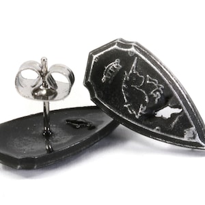 May include: A pair of black shield-shaped earrings with a silver-toned post and back. The shield has a design of a unicorn with a crown on its head.