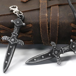 May include: Two silver-toned dagger pendants, one on a black cord and the other on a silver key chain. Both pendants feature intricate details and a pointed tip.