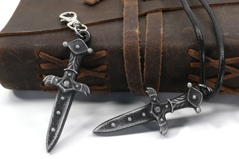 May include: Two silver-toned dagger pendants, one on a black cord and the other on a silver key chain. Both pendants feature intricate details and a pointed tip.