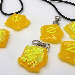 May include: Five yellow resin charms with a black and white symbol on each. The charms are on black cords and silver chains. Some charms have glitter embedded in the resin.