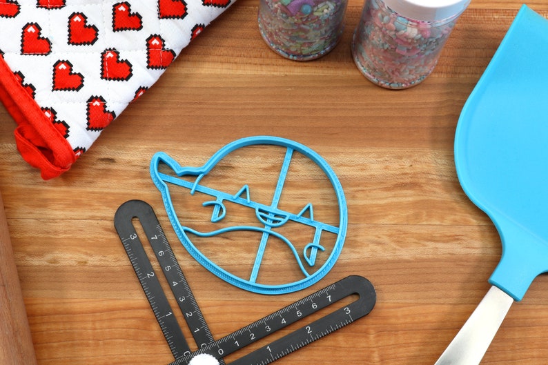 Peut inclure: Emporte-pi&egrave;ce en plastique bleu en forme de fant&ocirc;me avec un visage souriant. L'emporte-pi&egrave;ce est sur une surface en bois avec d'autres accessoires de p&acirc;tisserie.