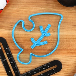 May include: A blue plastic cookie cutter shaped like a leaf with a small branch inside. The cutter is on a wooden surface.