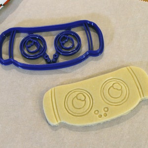 May include: A blue plastic cookie cutter and a cut-out cookie. The cookie cutter has a rectangular shape with two circular designs. The cut-out cookie has the same design, with two circular impressions and small dots.