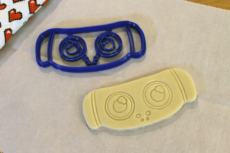 May include: A blue plastic cookie cutter and a cut-out cookie. The cookie cutter has a rectangular shape with two circular designs. The cut-out cookie has the same design, with two circular impressions and small dots.