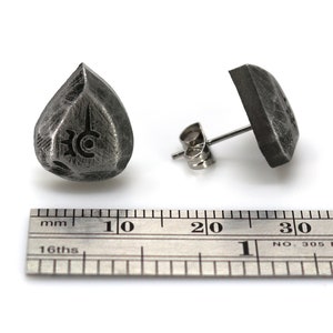 May include: A pair of silver stud earrings, one shaped like a teardrop with a symbol engraved on it, and the other a square shape. The earrings are shown next to a ruler for size reference.