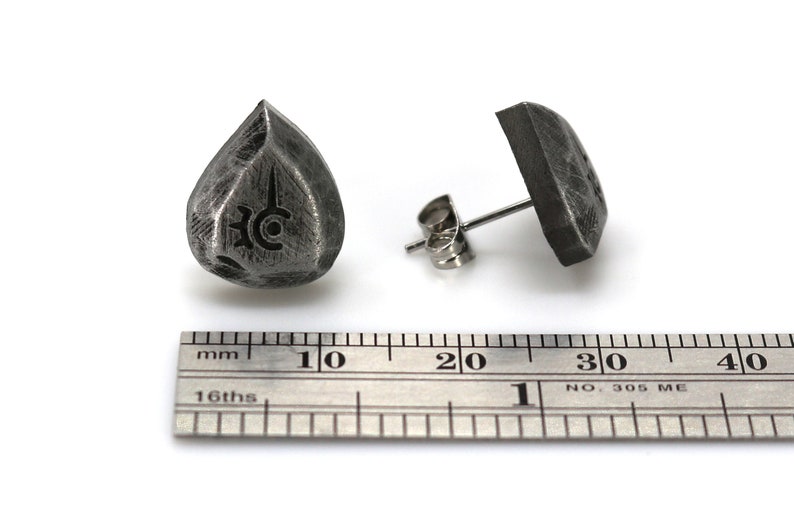 May include: A pair of silver stud earrings, one shaped like a teardrop with a symbol engraved on it, and the other a square shape. The earrings are shown next to a ruler for size reference.
