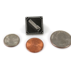 May include: A silver-toned metal pin with a black square background and a white image of a paint can with a brush. The pin is shown next to three US coins: a quarter, a dime, and a penny.