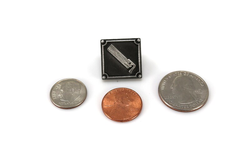 May include: A silver-toned metal pin with a black square background and a white image of a paint can with a brush. The pin is shown next to three US coins: a quarter, a dime, and a penny.