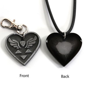 Puede incluir: Un colgante de coraz&oacute;n plateado con un coraz&oacute;n alado y un dise&ntilde;o de tri&aacute;ngulo en la parte delantera. La parte posterior del colgante es negra y lisa. El colgante est&aacute; unido a un cord&oacute;n negro.