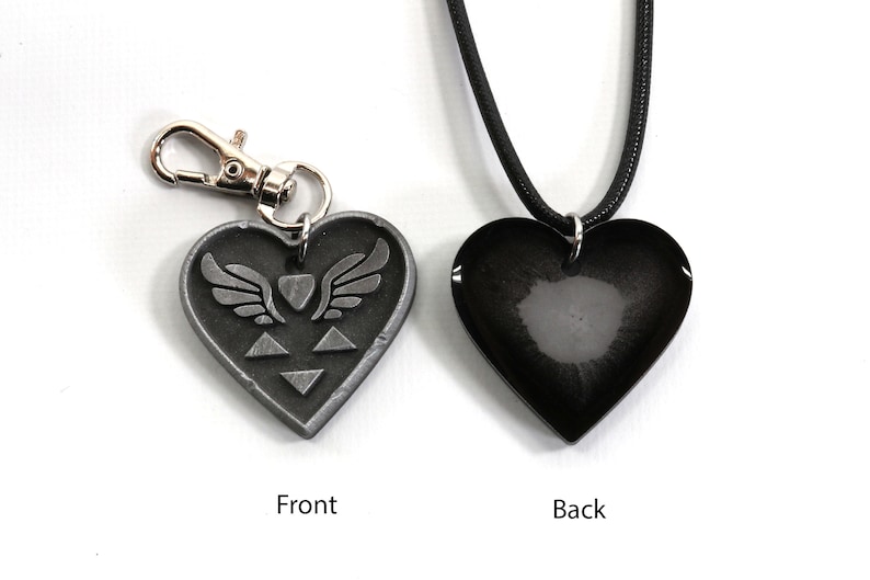 Puede incluir: Un colgante de coraz&oacute;n plateado con un coraz&oacute;n alado y un dise&ntilde;o de tri&aacute;ngulo en la parte delantera. La parte posterior del colgante es negra y lisa. El colgante est&aacute; unido a un cord&oacute;n negro.