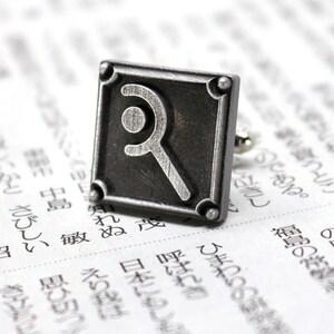 May include: A square, silver-toned cufflink with a stylized letter "R" design. The cufflink has a textured border and is set against a backdrop of printed text, suggesting a literary or intellectual theme.
