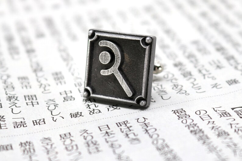 May include: A square, silver-toned cufflink with a stylized letter "R" design. The cufflink has a textured border and is set against a backdrop of printed text, suggesting a literary or intellectual theme.