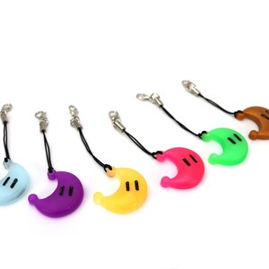 May include: Six colorful crescent moon-shaped keychains with black eyes. The keychains are attached to black cords with silver clasps. The colors of the keychains are light blue, purple, yellow, pink, green, and brown.