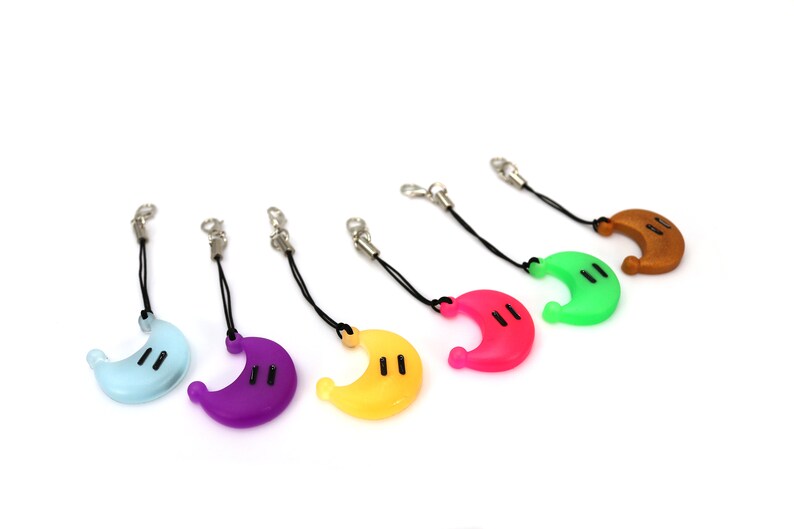 May include: Six colorful crescent moon-shaped keychains with black eyes. The keychains are attached to black cords with silver clasps. The colors of the keychains are light blue, purple, yellow, pink, green, and brown.
