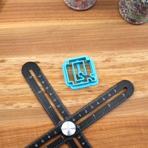 May include: A blue plastic cookie cutter shaped like the letter Q, on a wooden surface with a black ruler.