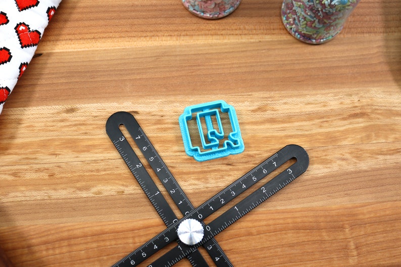 May include: A blue plastic cookie cutter shaped like the letter Q, on a wooden surface with a black ruler.