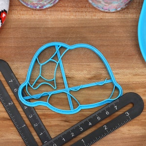 May include: A blue plastic cookie cutter shaped like a turtle shell. The cutter is about 3 inches wide and 2 inches tall. It is sitting on a wooden surface with a ruler next to it.