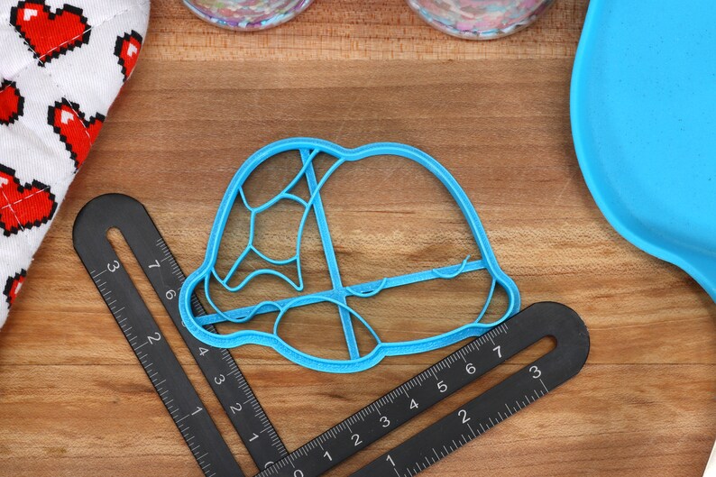 May include: A blue plastic cookie cutter shaped like a turtle shell. The cutter is about 3 inches wide and 2 inches tall. It is sitting on a wooden surface with a ruler next to it.