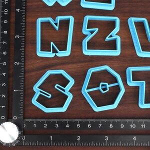 May include: A set of blue plastic cookie cutters shaped like the letters N, Z, V, S, O, and T. The cutters are arranged on a wooden surface with a ruler.
