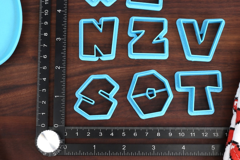 May include: A set of blue plastic cookie cutters shaped like the letters N, Z, V, S, O, and T. The cutters are arranged on a wooden surface with a ruler.