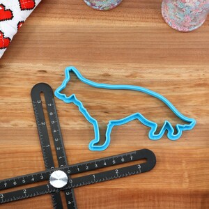 May include: A blue plastic cookie cutter shaped like a German Shepherd dog on a wooden surface with a black measuring tool.