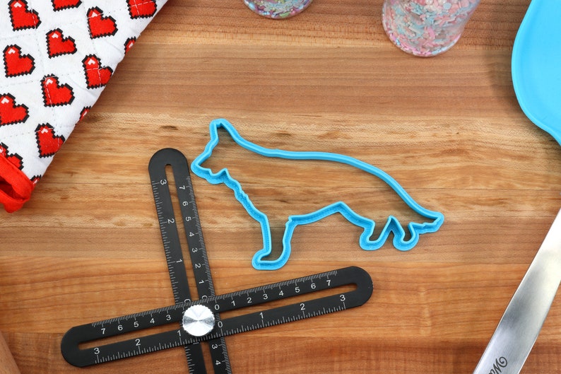 May include: A blue plastic cookie cutter shaped like a German Shepherd dog on a wooden surface with a black measuring tool.