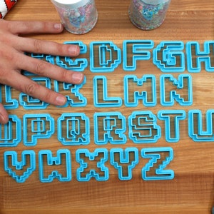 May include: A set of blue plastic cookie cutters shaped like the letters of the alphabet in a pixelated style. The cutters are arranged in alphabetical order on a wooden surface.