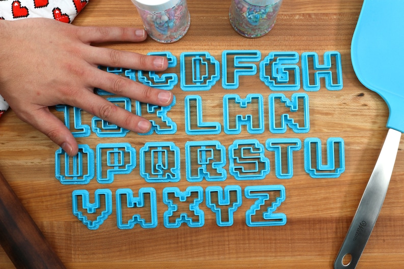 May include: A set of blue plastic cookie cutters shaped like the letters of the alphabet in a pixelated style. The cutters are arranged in alphabetical order on a wooden surface.