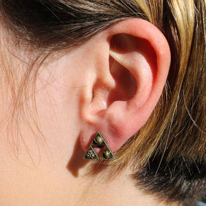 May include: A close-up of a person's ear with a gold triangle earring. The earring has a geometric design with multiple small circles.