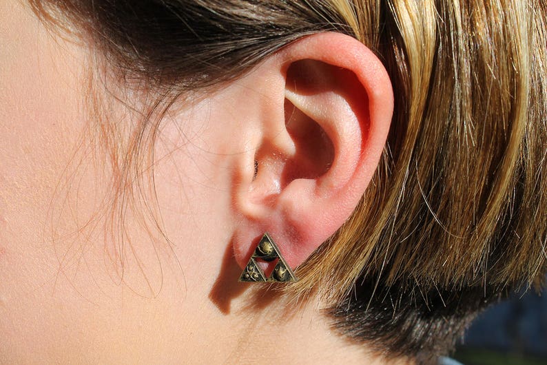 May include: A close-up of a person's ear with a gold triangle earring. The earring has a geometric design with multiple small circles.