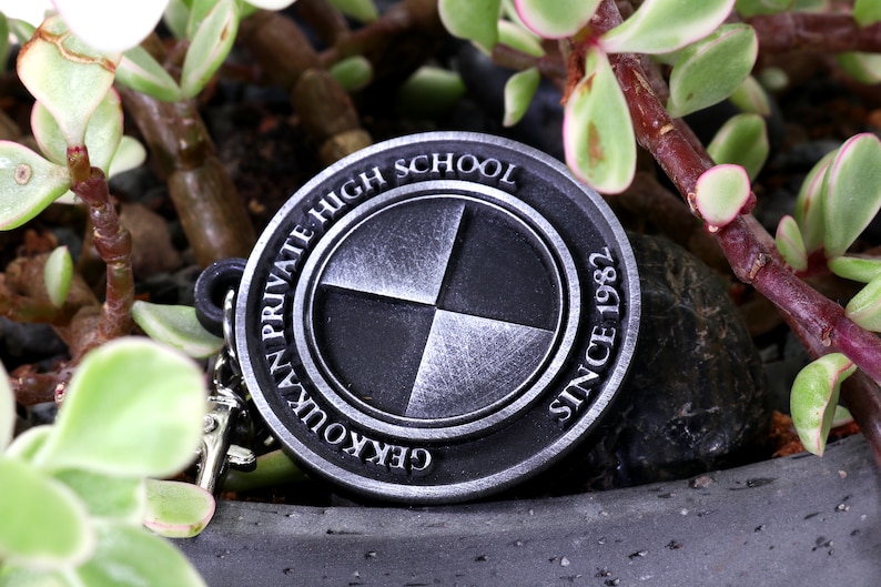 May include: A silver-colored metal keychain with the words "Gekkoukan Private High School Since 1982" around the edge. The center of the keychain features a four-quadrant design.