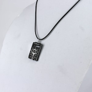 May include: A black necklace with a silver pendant featuring a stylised eye design. The pendant is rectangular and has a textured surface.