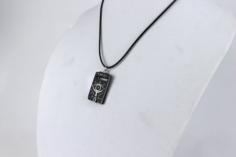 May include: A black necklace with a silver pendant featuring a stylised eye design. The pendant is rectangular and has a textured surface.