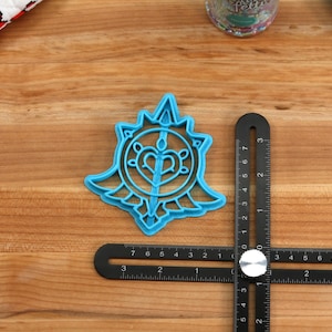 May include: A blue plastic cookie cutter shaped like a stylized heart with wings and a sword inside. The cutter is on a wooden surface with a ruler.