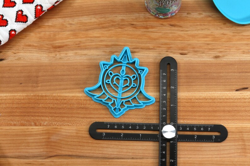 May include: A blue plastic cookie cutter shaped like a stylized heart with wings and a sword inside. The cutter is on a wooden surface with a ruler.