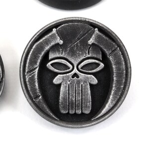May include: A silver-colored metal token with a skull design. The skull has two large eyes and a detailed jawline. The skull is surrounded by a circular border with a textured surface.