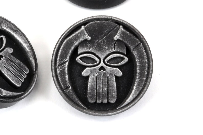 May include: A silver-colored metal token with a skull design. The skull has two large eyes and a detailed jawline. The skull is surrounded by a circular border with a textured surface.