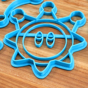 May include: A set of blue plastic cookie cutters in the shape of a sun, a circle, a crescent moon, and a rectangle. The sun cookie cutter has a face with two eyes.