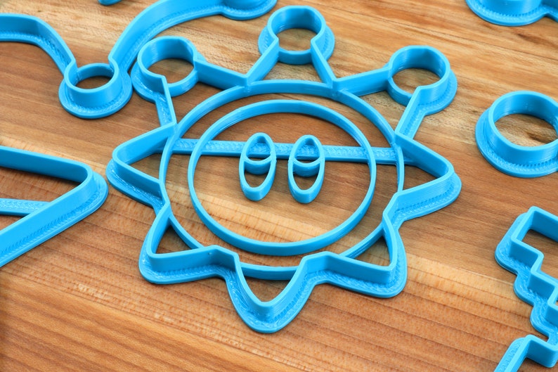 May include: A set of blue plastic cookie cutters in the shape of a sun, a circle, a crescent moon, and a rectangle. The sun cookie cutter has a face with two eyes.