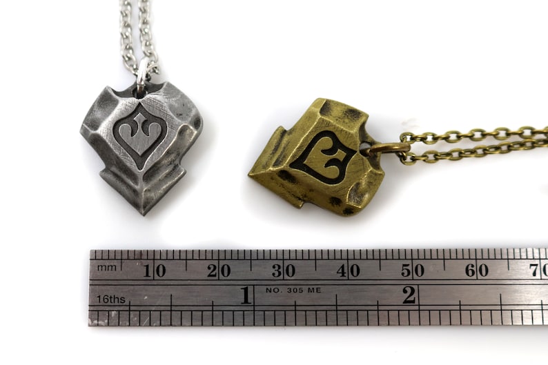 Pode incluir: Dois pingentes, um prateado e outro dourado, com um design em forma de cora&ccedil;&atilde;o e uma r&eacute;gua para refer&ecirc;ncia de tamanho. Os pingentes medem aproximadamente 3 cm de comprimento.