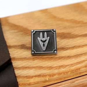 Op de afbeelding: Een zilveren metalen pin met een zwart en zilver ontwerp. De pin heeft een gestileerde, puntige vorm met drie punten. De pin is geplaatst tegen een houten achtergrond.