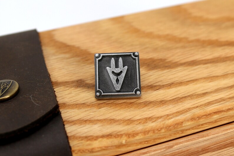 Op de afbeelding: Een zilveren metalen pin met een zwart en zilver ontwerp. De pin heeft een gestileerde, puntige vorm met drie punten. De pin is geplaatst tegen een houten achtergrond.