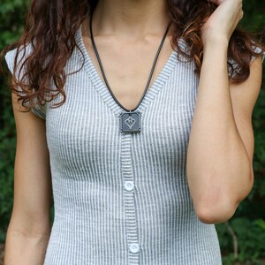 May include: A woman wearing a gray and white striped dress with a black necklace and a square pendant featuring a heart design.