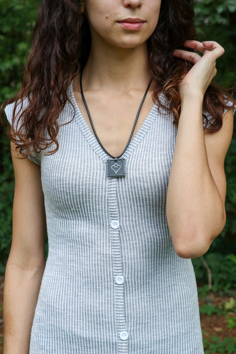 May include: A woman wearing a gray and white striped dress with a black necklace and a square pendant featuring a heart design.