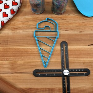 May include: Blue plastic ice cream cone cookie cutter on a wooden surface with a black ruler, a rolling pin, and a blue spatula.