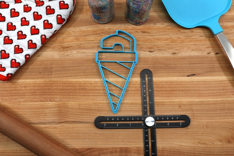 May include: Blue plastic ice cream cone cookie cutter on a wooden surface with a black ruler, a rolling pin, and a blue spatula.