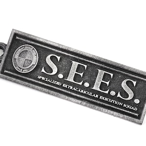 May include: A silver metal keychain with the text "S.E.E.S. SPECIALIZED EXTRACURRICULAR EXECUTION SQUAD" and a circular logo with a cross inside.