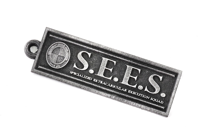 May include: A silver metal keychain with the text "S.E.E.S. SPECIALIZED EXTRACURRICULAR EXECUTION SQUAD" and a circular logo with a cross inside.