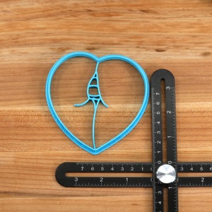 May include: A blue plastic heart-shaped cookie cutter with a design inside, measured against a black ruler with a silver knob.