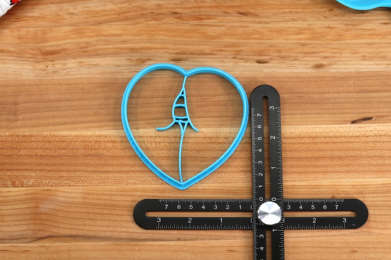 May include: A blue plastic heart-shaped cookie cutter with a design inside, measured against a black ruler with a silver knob.